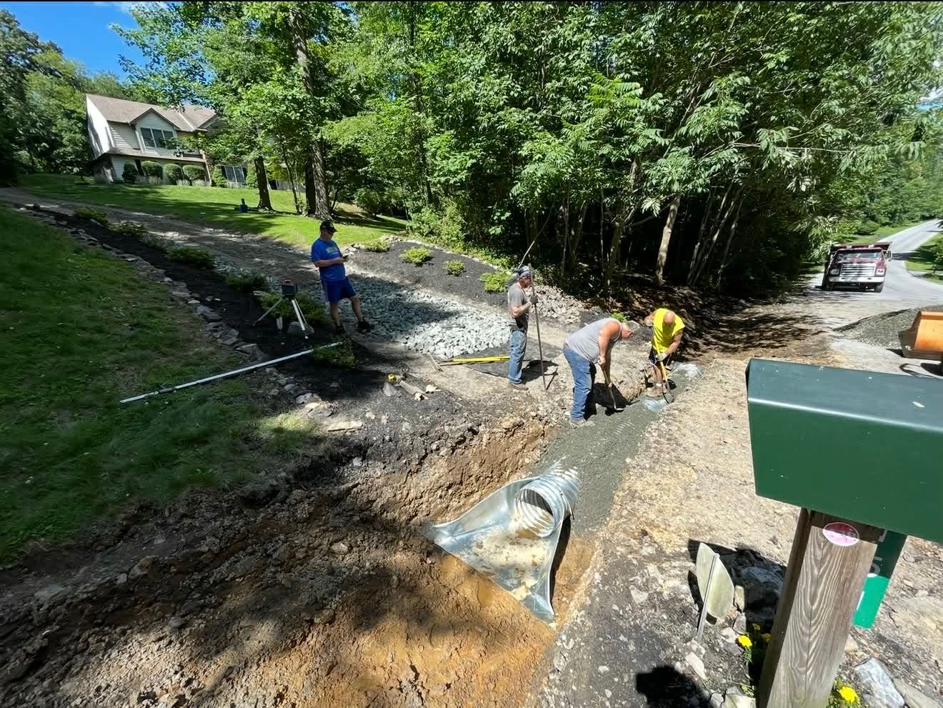 Workers installing a gravel path on a wooded hillside beside a road and houses