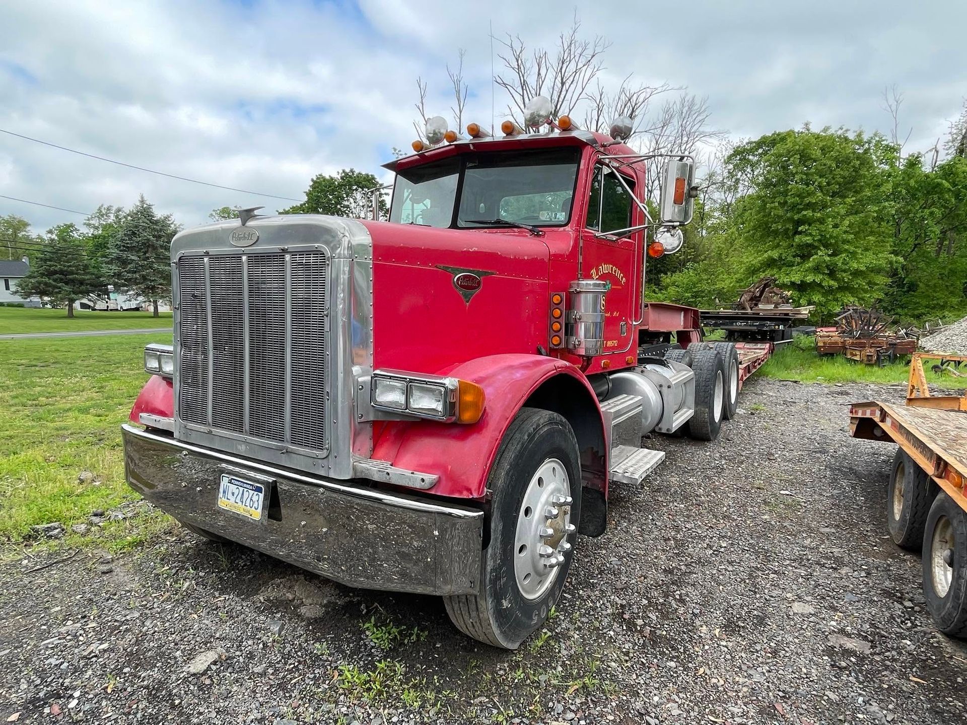Red semi truck parked on gravel under cloudy sky, with a chrome grille and flatbed trailer.