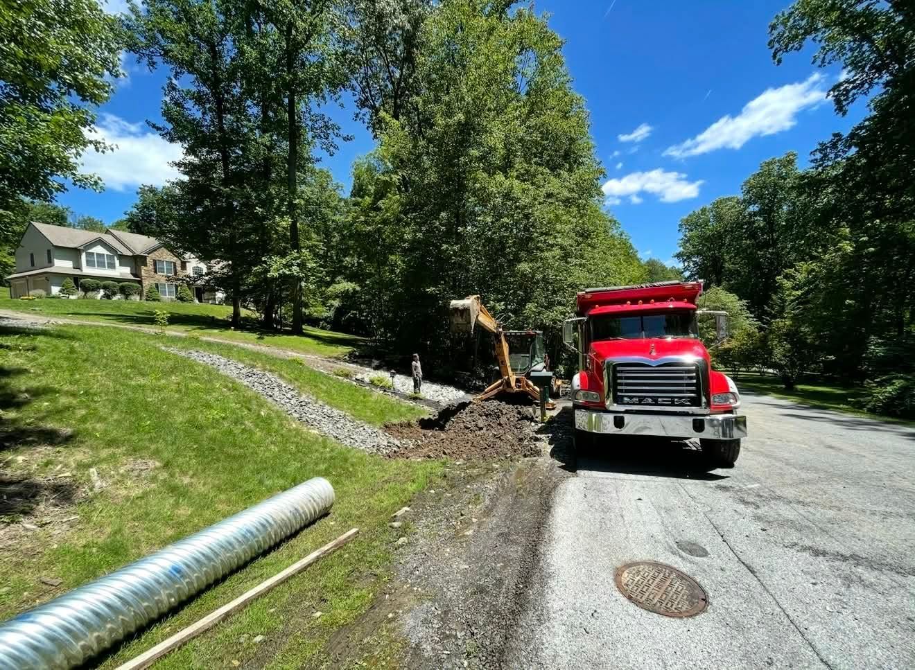 Red dump truck beside roadside excavation with mud and a large pipe on a sunny tree-lined street