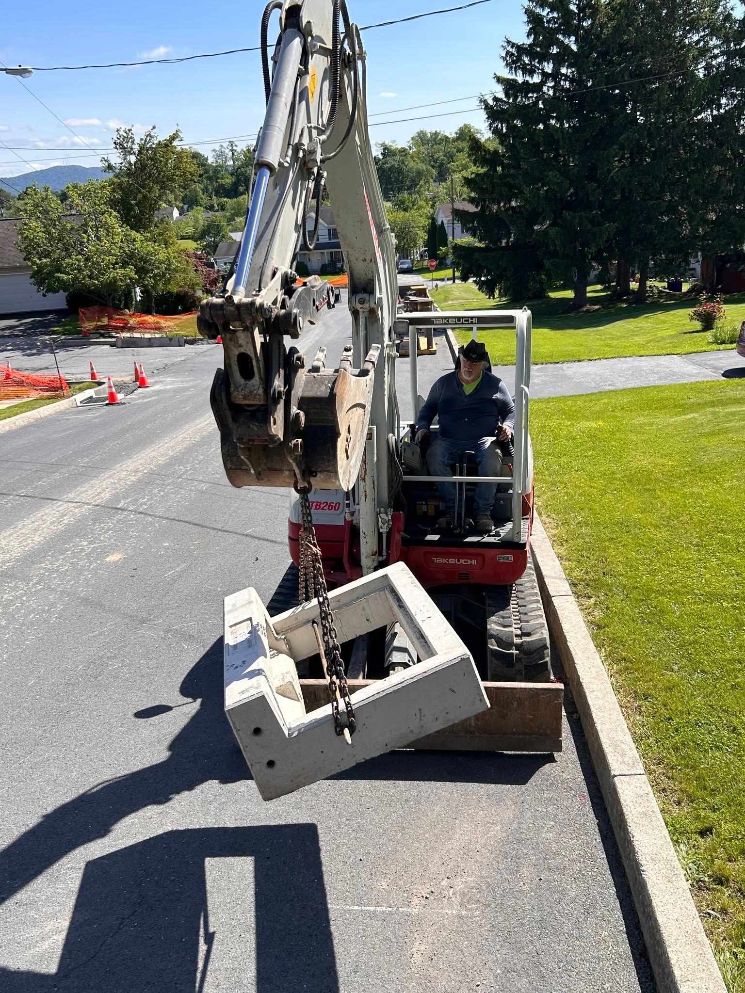 Backhoe parked on roadside beside grass, with a worker in the cab and a concrete box in front
