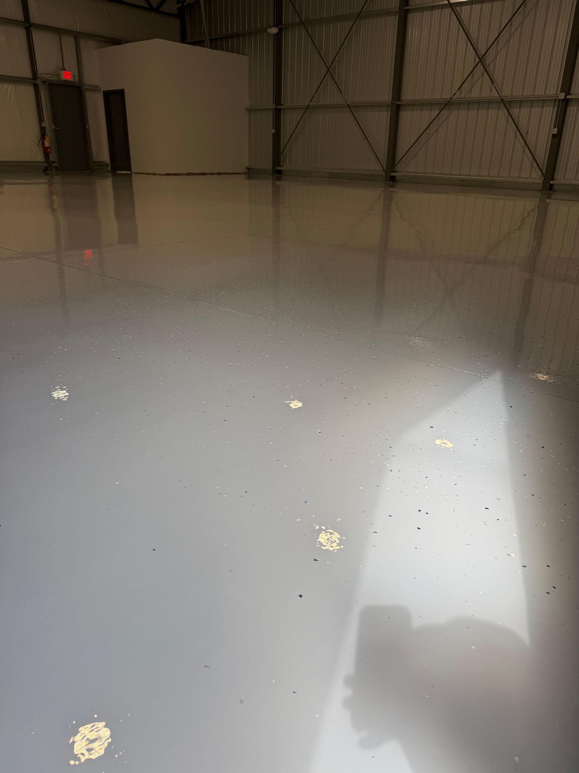 Glossy gray warehouse floor reflecting lights and walls, with scattered debris and a shadow in the foreground