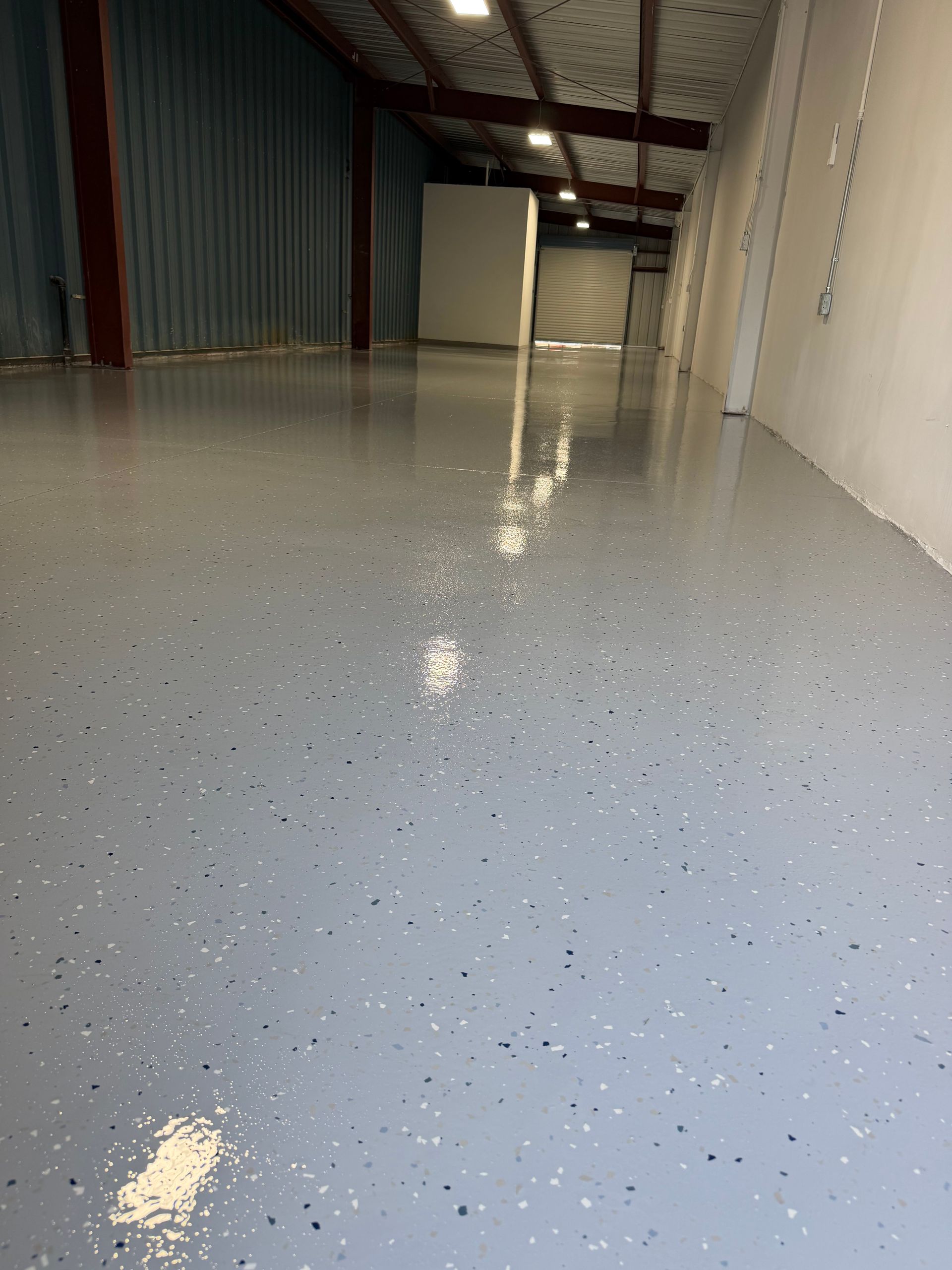 Empty warehouse corridor with glossy speckled gray floor and white walls under overhead lights