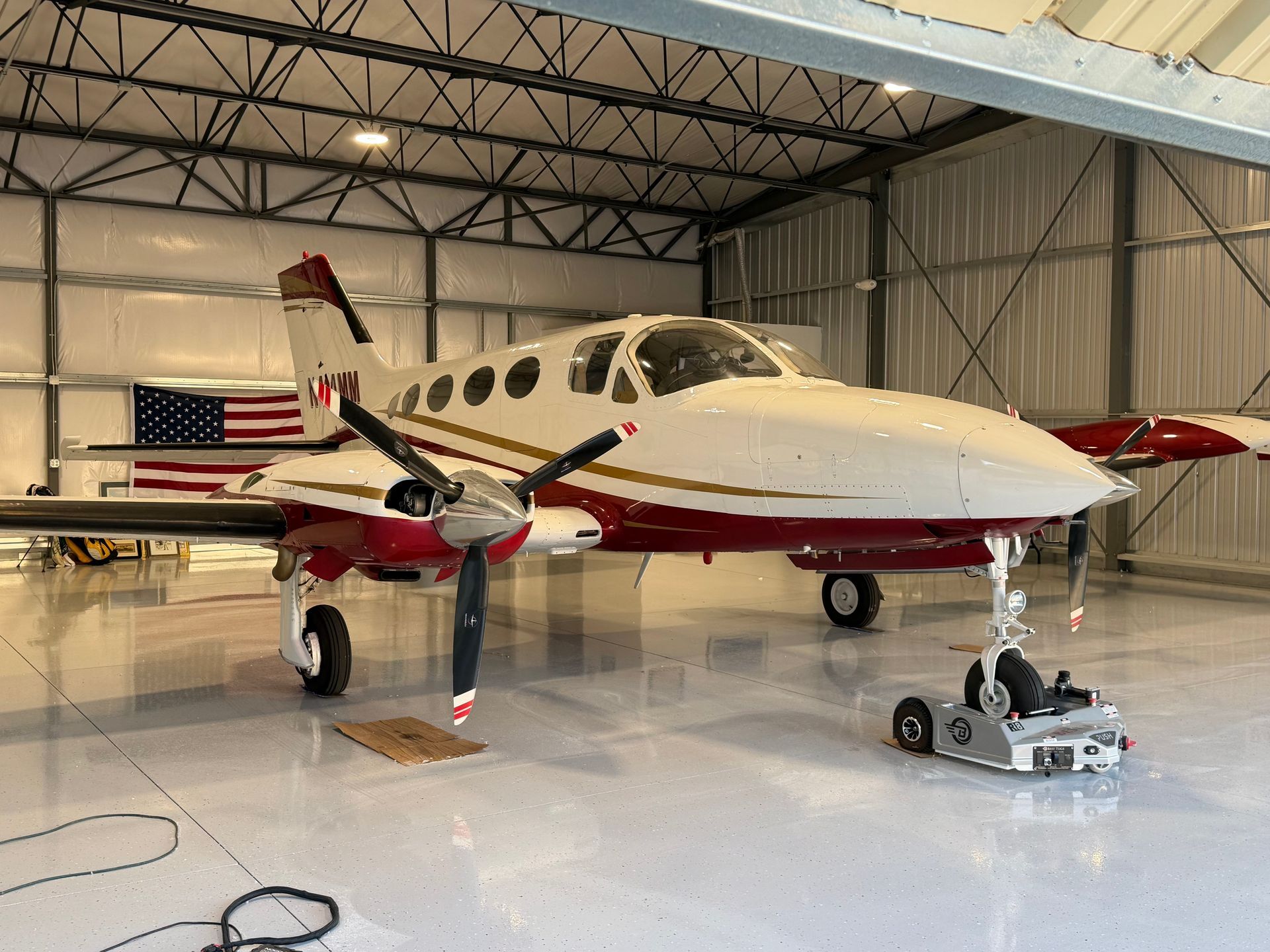 Small twin-engine prop plane in a hangar, white with red accents, on landing gear with maintenance equipment nearby