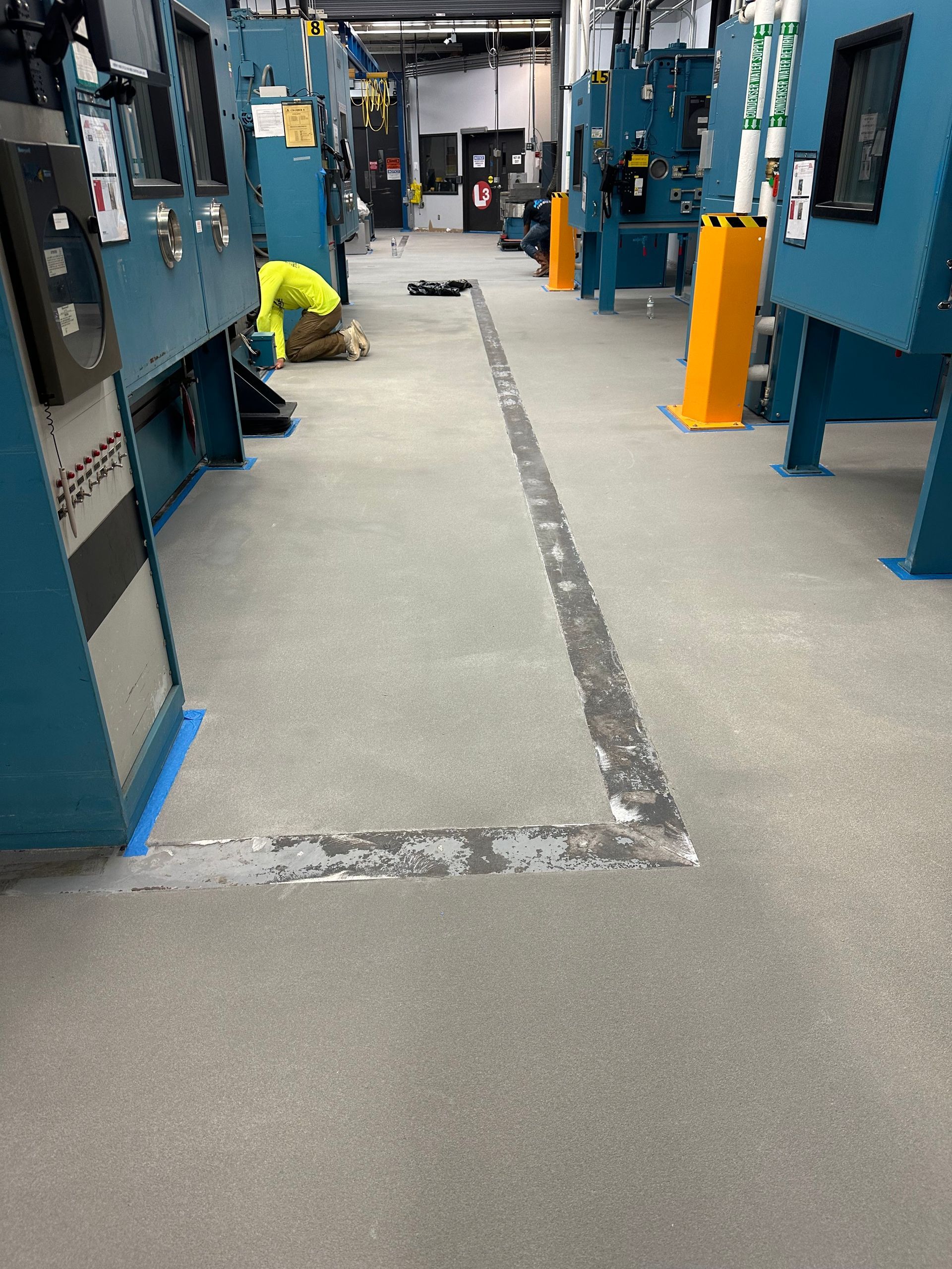 Industrial room with blue machinery and a marked walkway; a worker kneels in the background.
