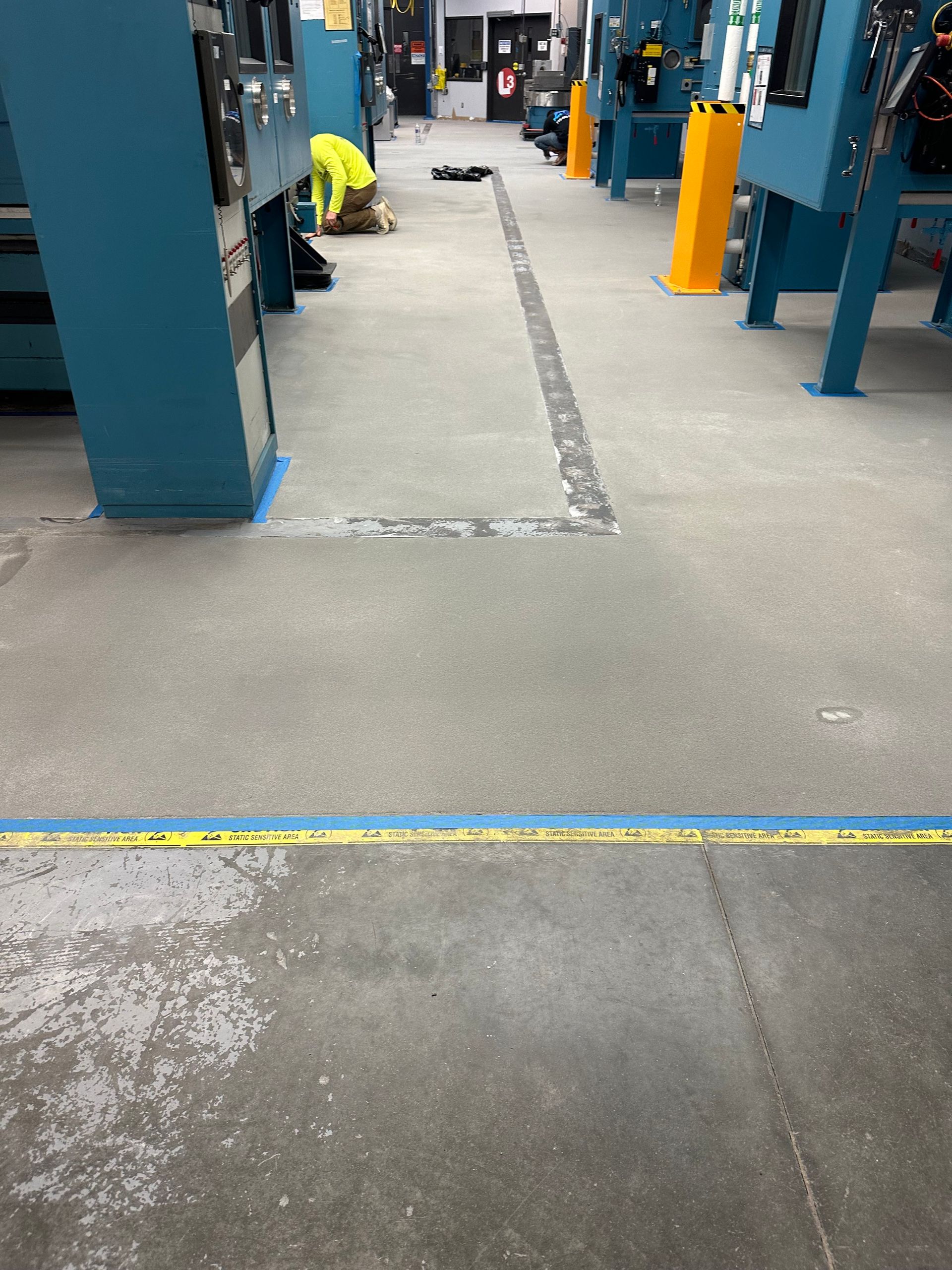 Industrial floor with a long cracked repair seam between blue columns and yellow posts, worker kneeling in background