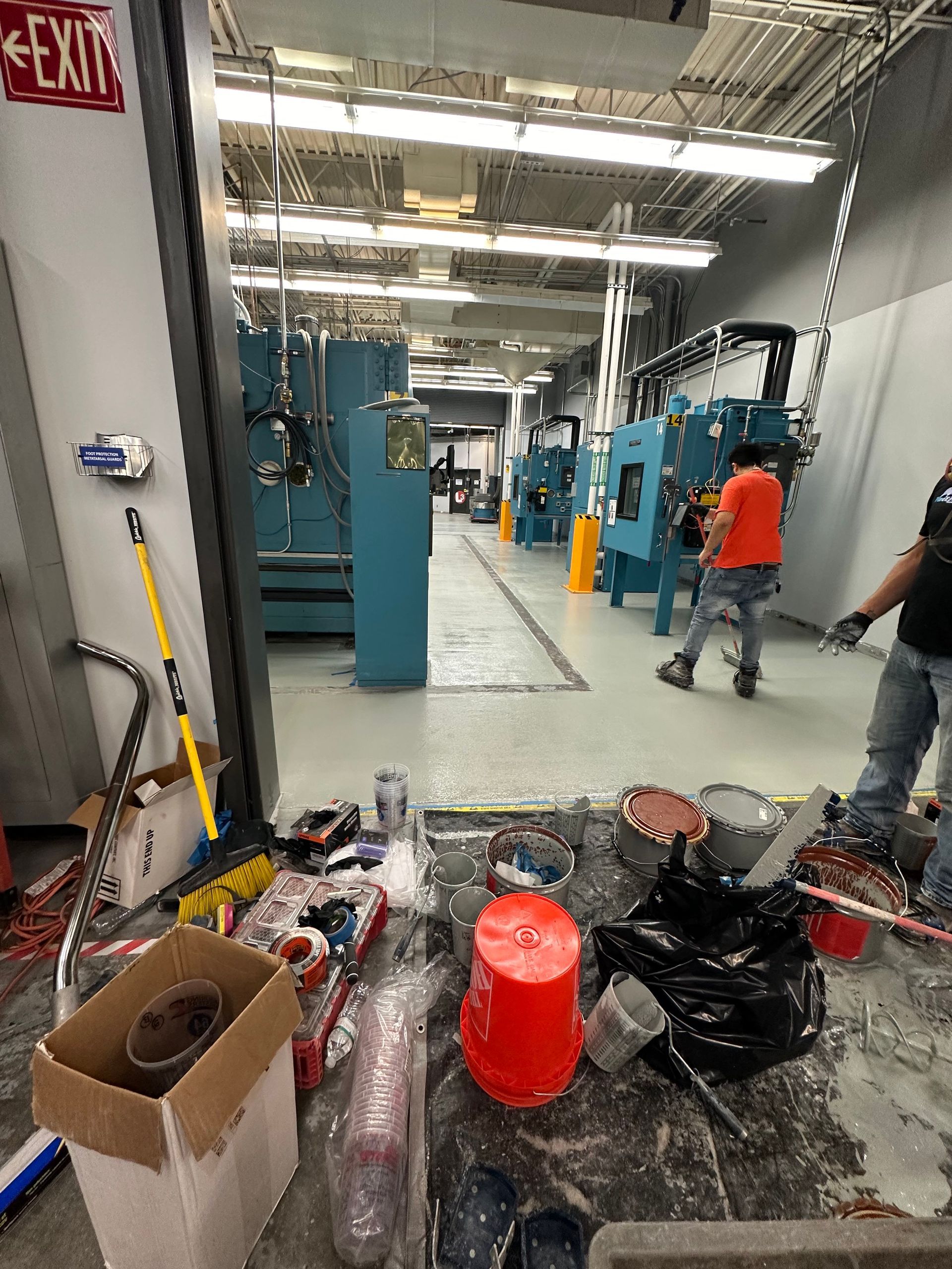 Industrial workshop aisle with scattered broken parts, orange bucket, and workers in the background