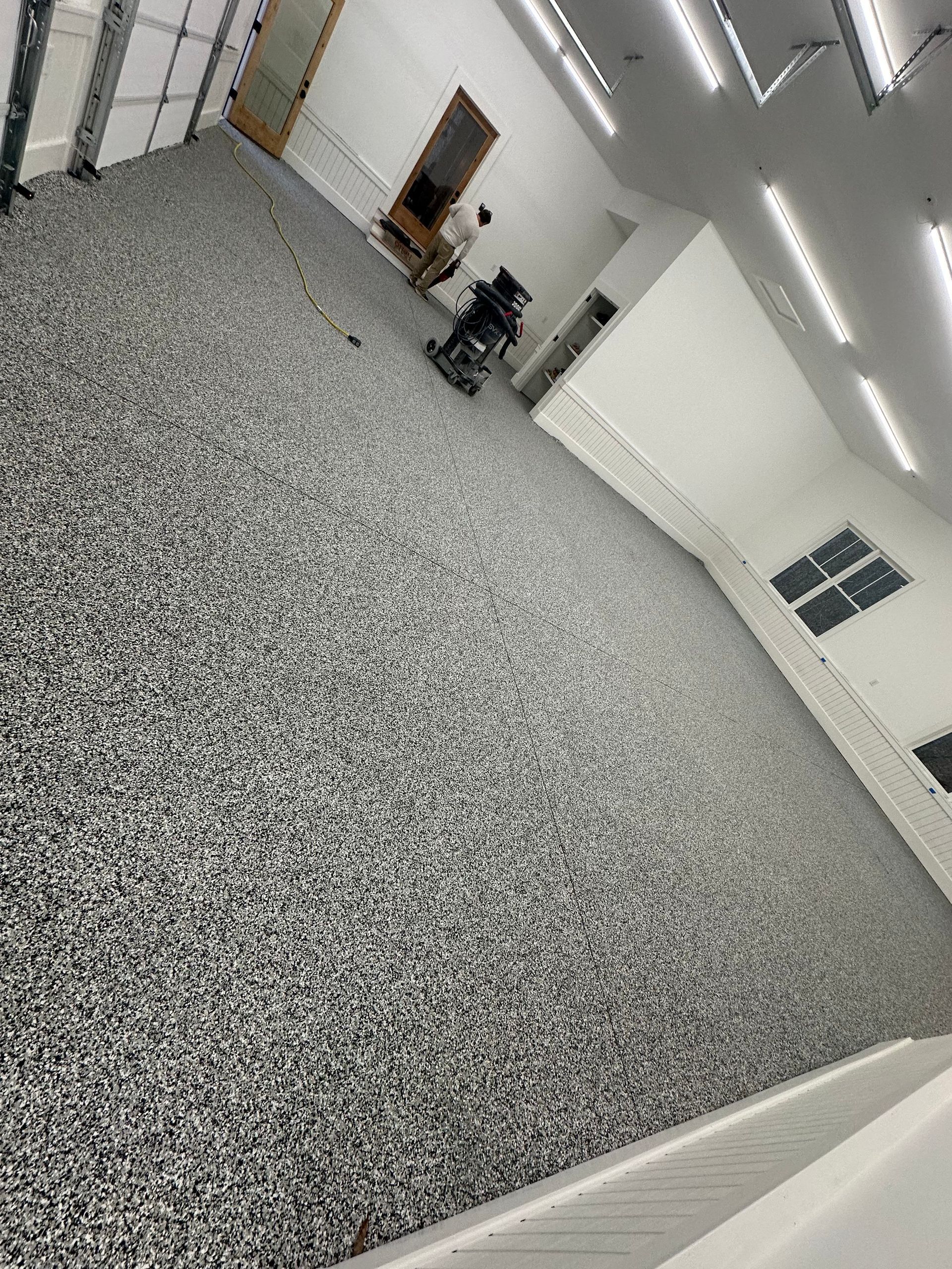 Empty indoor room with speckled gray floor, white walls, fluorescent lights, and a person near a doorway