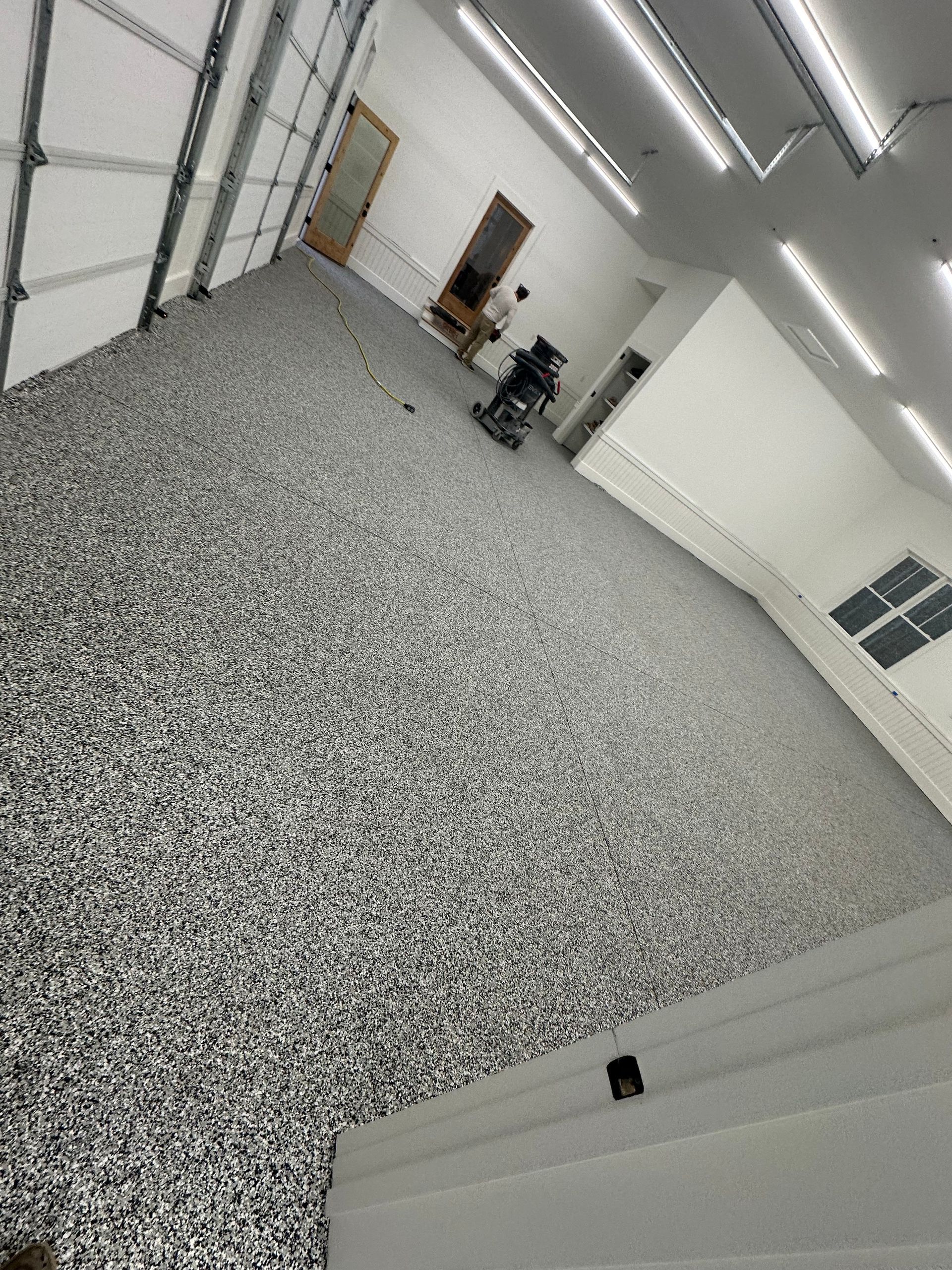Empty white garage-like room with speckled gray floor and overhead strip lights, viewed at an angle