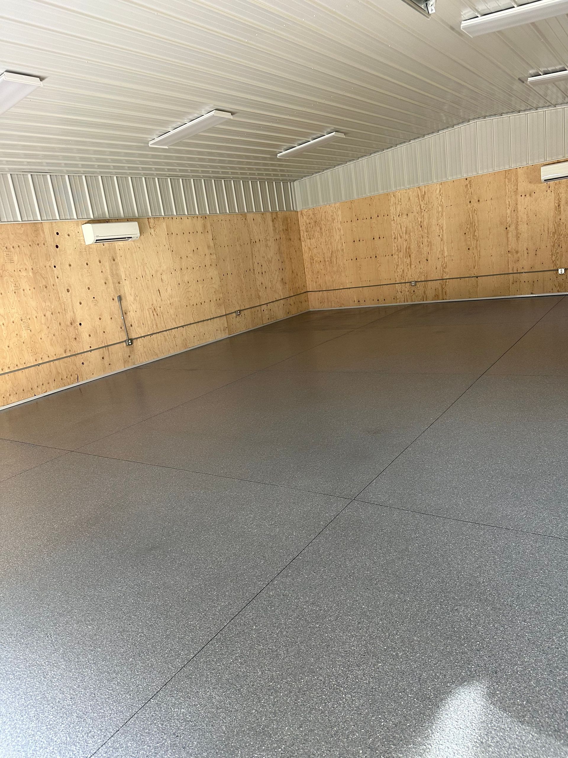 Empty insulated garage with plywood walls and gray speckled floor, lit by ceiling lights