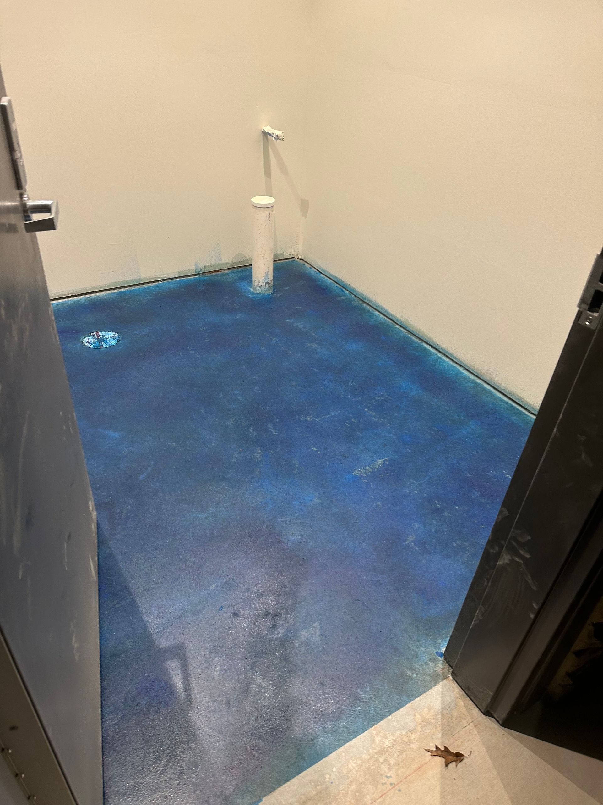 Small blue-floored utility room with beige walls, a toilet pipe in the corner, viewed through an open doorway