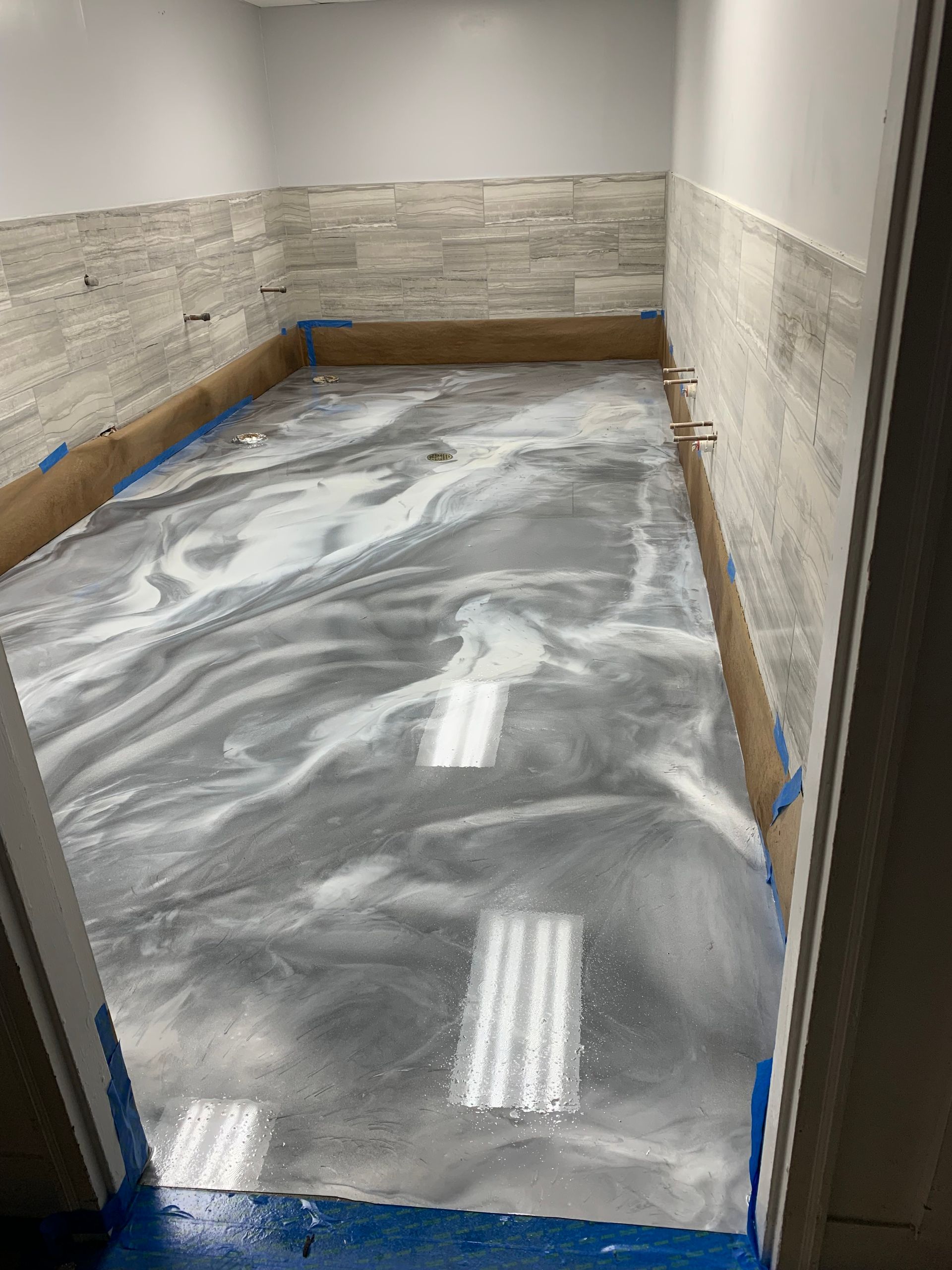 Empty hallway under renovation with wet gray floor coating and beige stone walls, masked with blue tape.