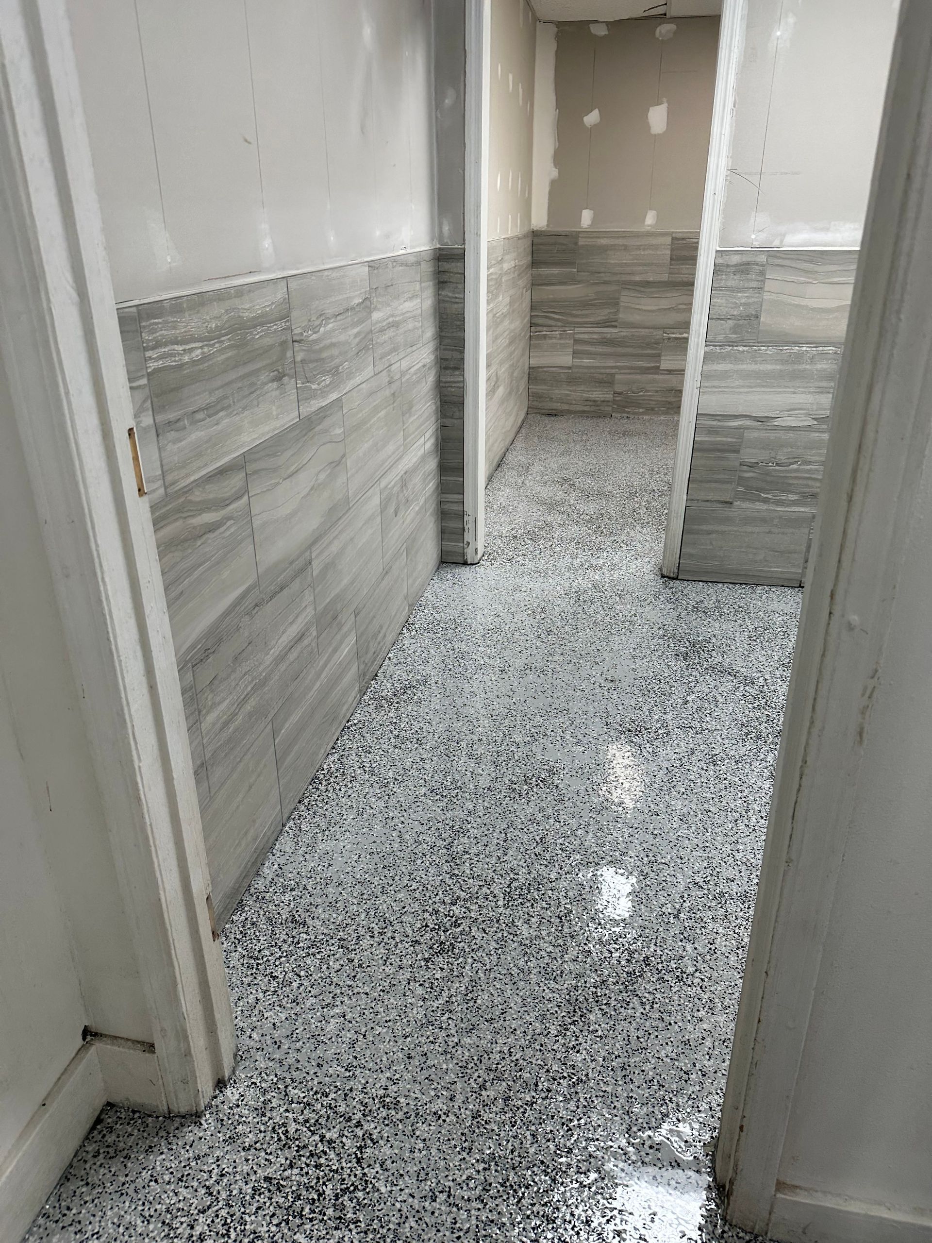 Narrow unfinished hallway with white walls and gray-speckled tile floor, leading to a doorway.