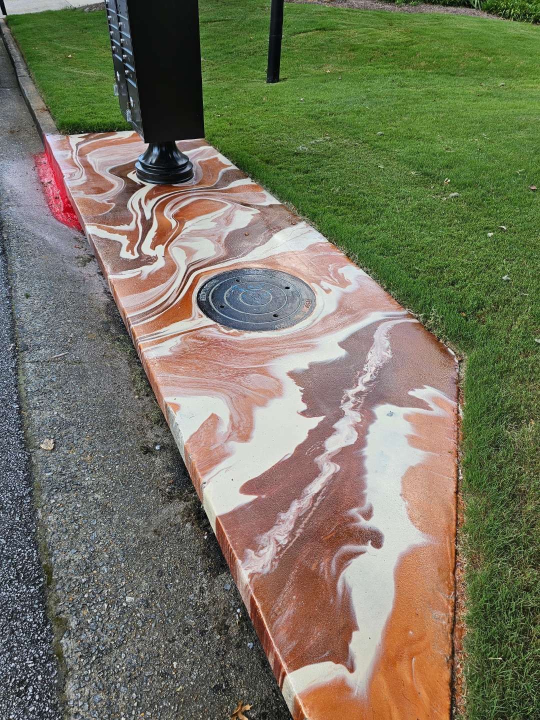 Marble-patterned painted curb beside grass and asphalt, with a black pole and round drain cover.