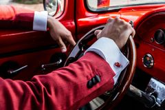 Een persoon in een rood, gestructureerd colbert met witte manchetten houdt het houten stuurwiel vast van een rode oldtimer.