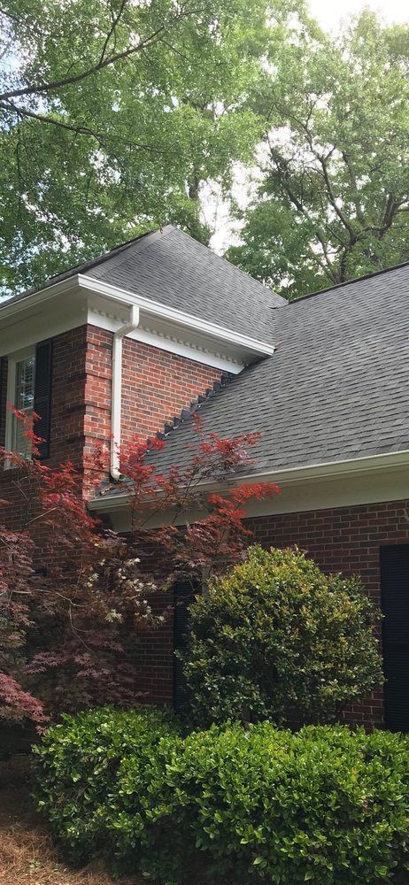 A brick house with a black roof and a white trim surrounded by trees and bushes.