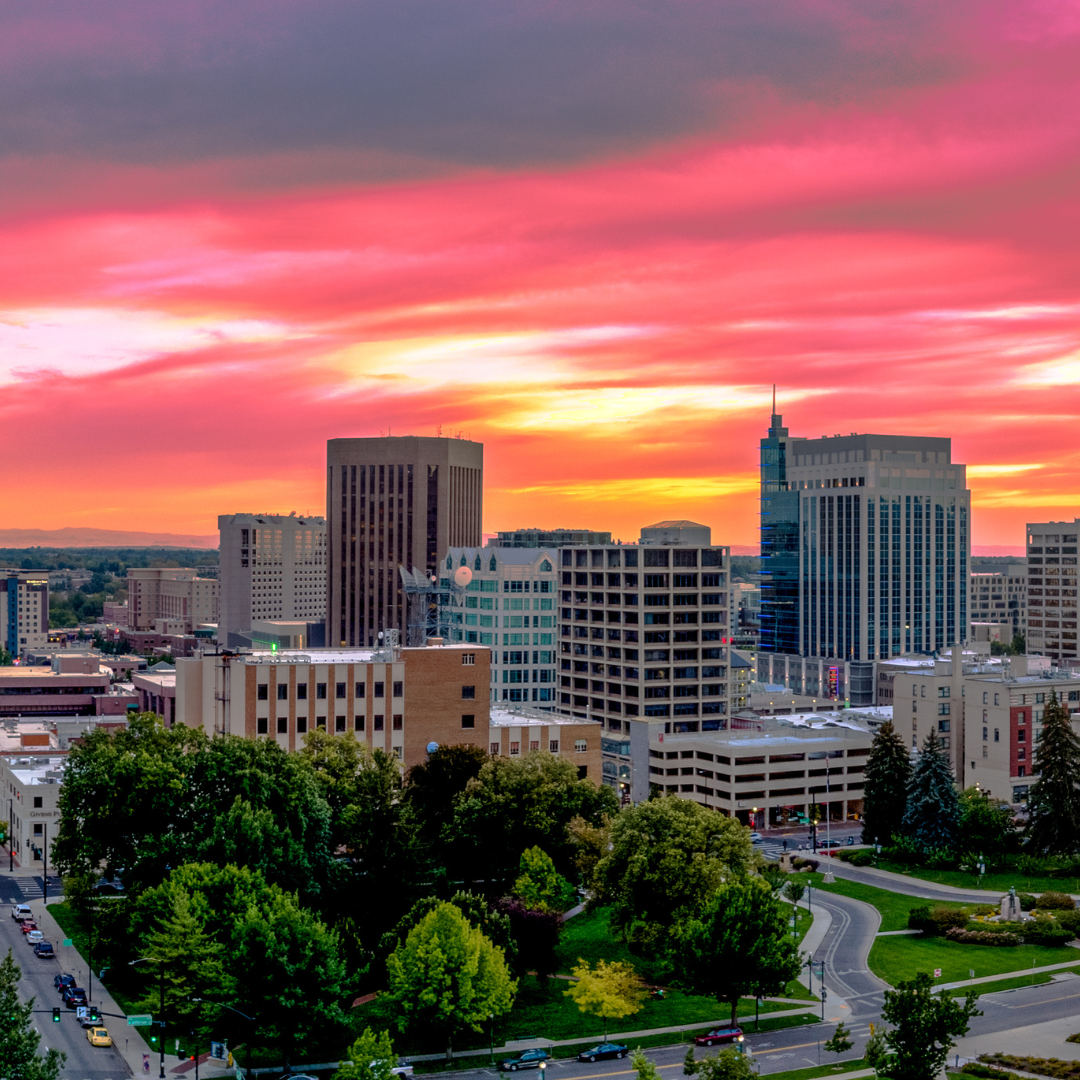 Boise, Idaho skyline at sunset, pink and orange sky over city buildings and green trees.