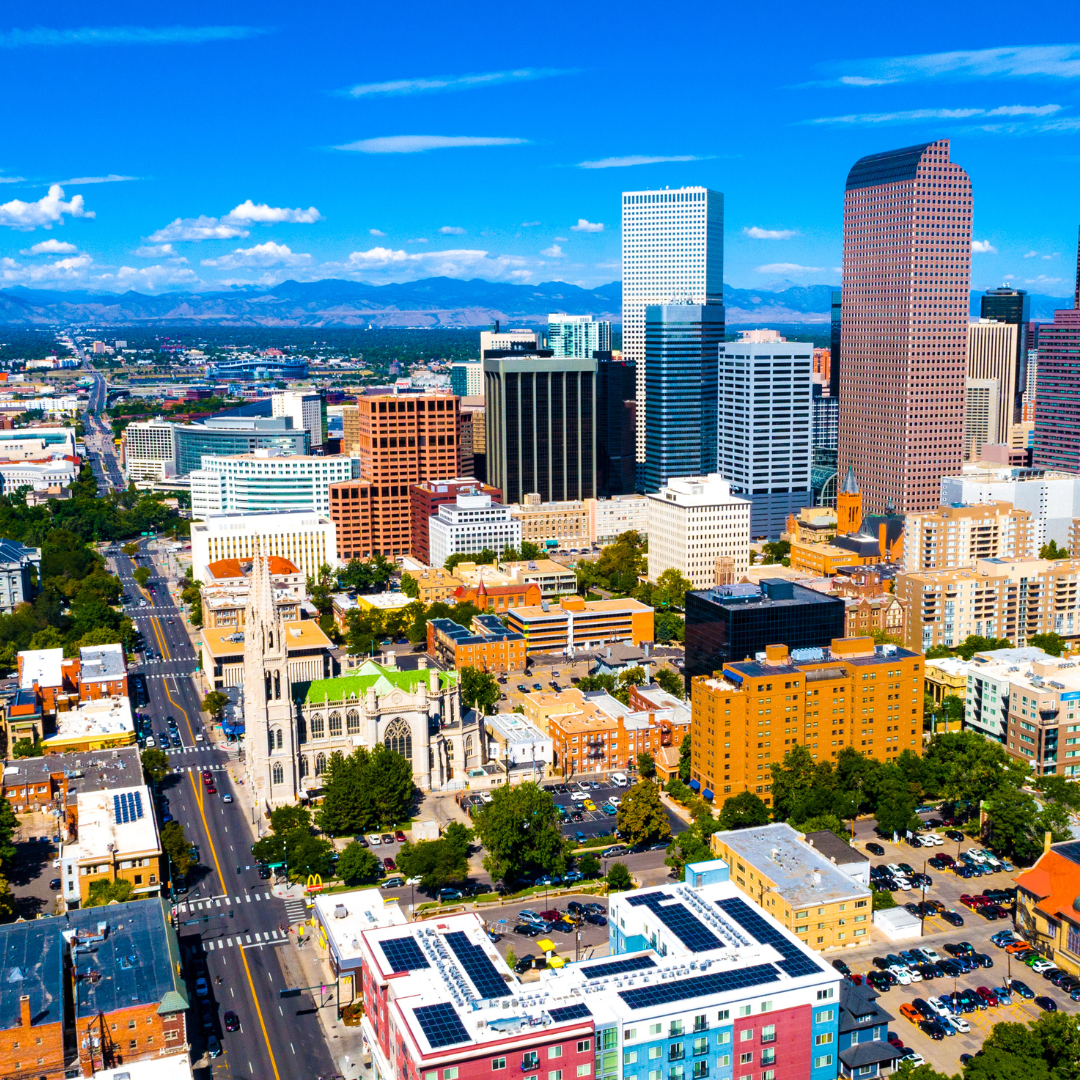 Cityscape of Denver, Colorado with tall buildings under a blue sky, mountains in the distance.