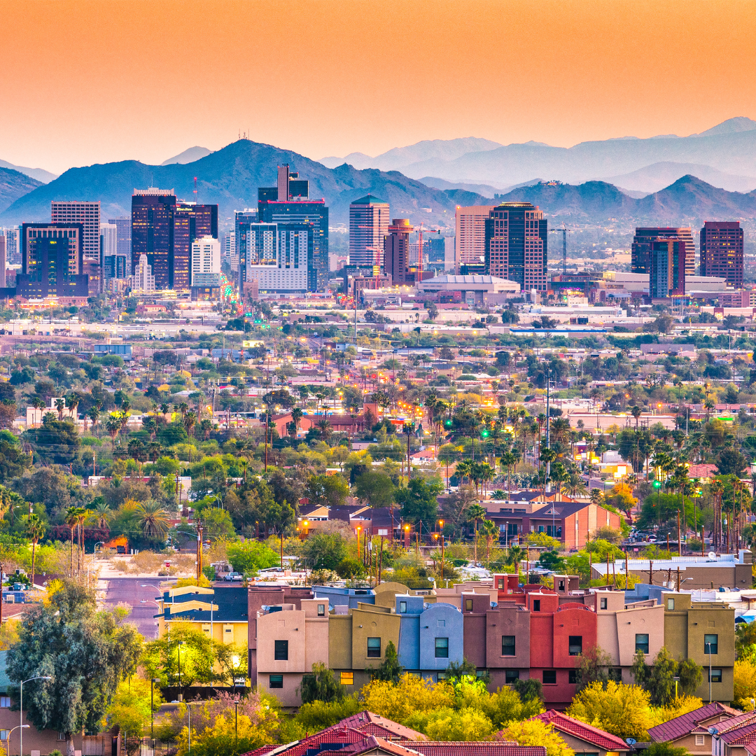 Phoenix, Arizona skyline with buildings, surrounded by green trees and distant mountains, under a colorful sky.
