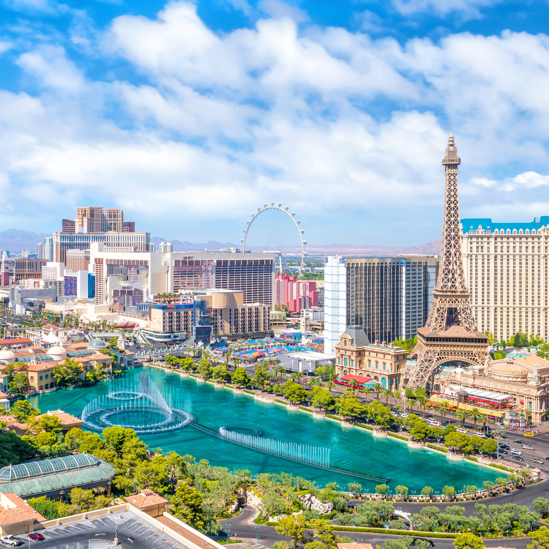 Las Vegas Strip with hotels, Eiffel Tower replica, water fountain show, and Ferris wheel under a blue sky.