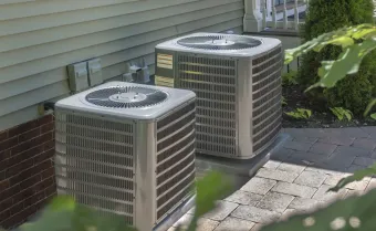 Two beige outdoor air conditioning units sit side-by-side next to a house with tan siding and brick trim.
