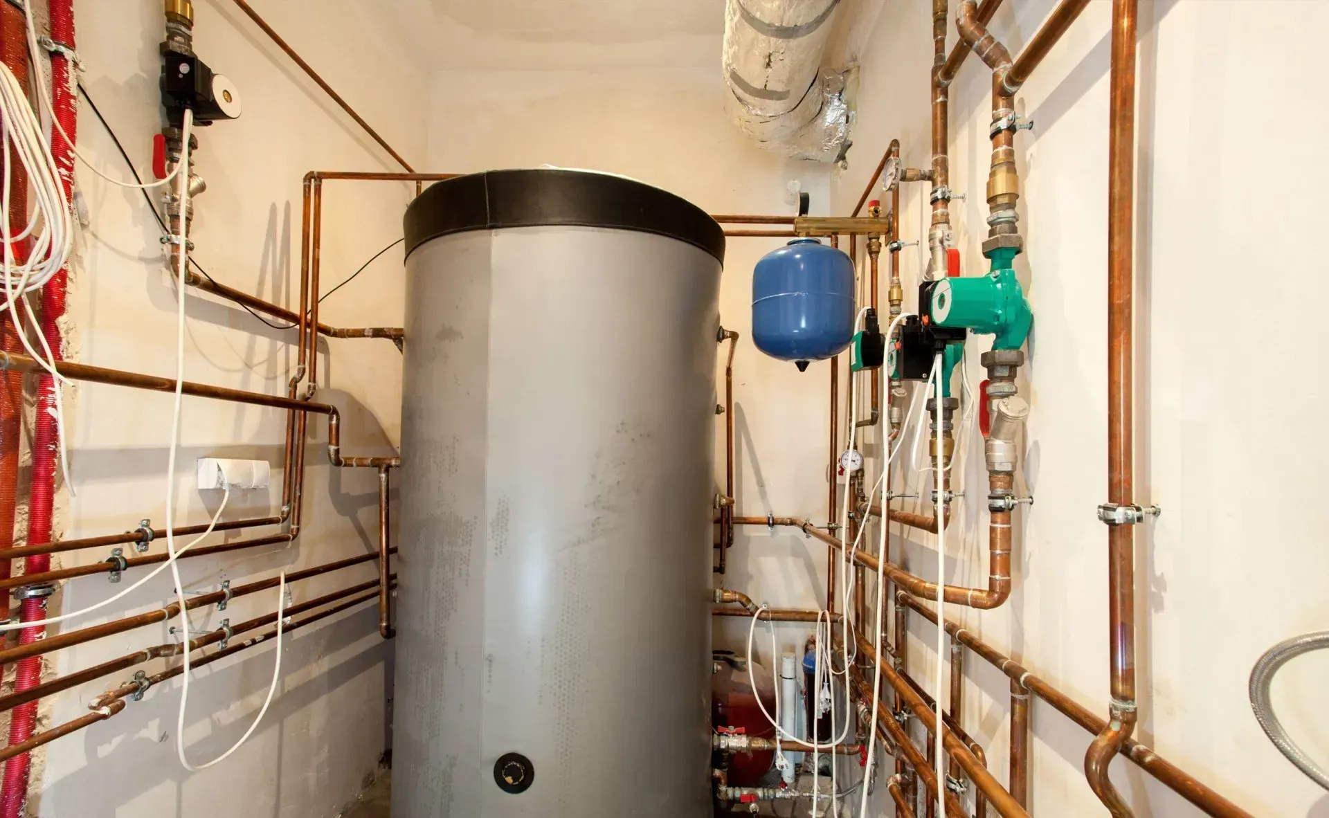 A grey hot water cylinder in a plant room, surrounded by a complex network of copper plumbing pipes and control valves.