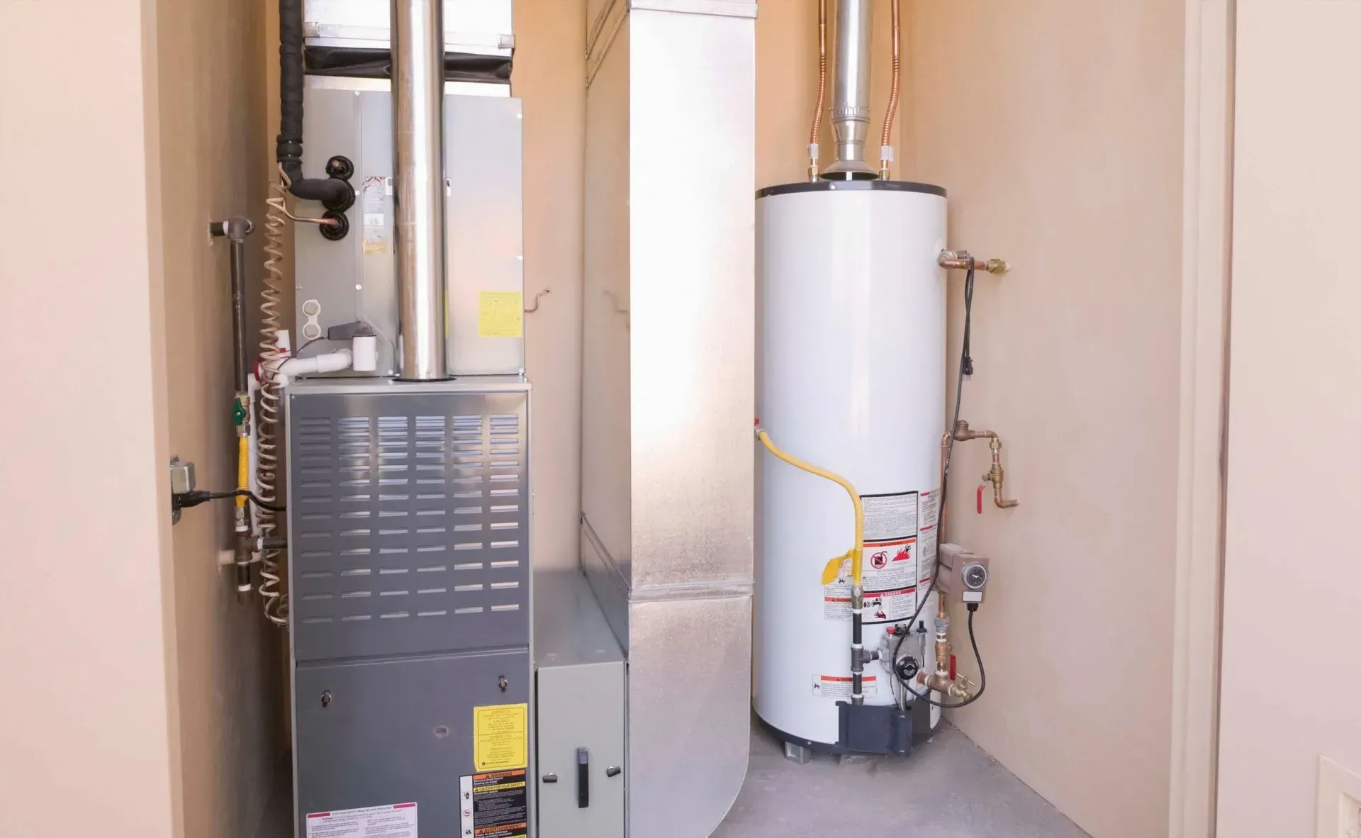 A grey furnace unit stands next to a tall white water heater inside a mechanical closet with beige walls.