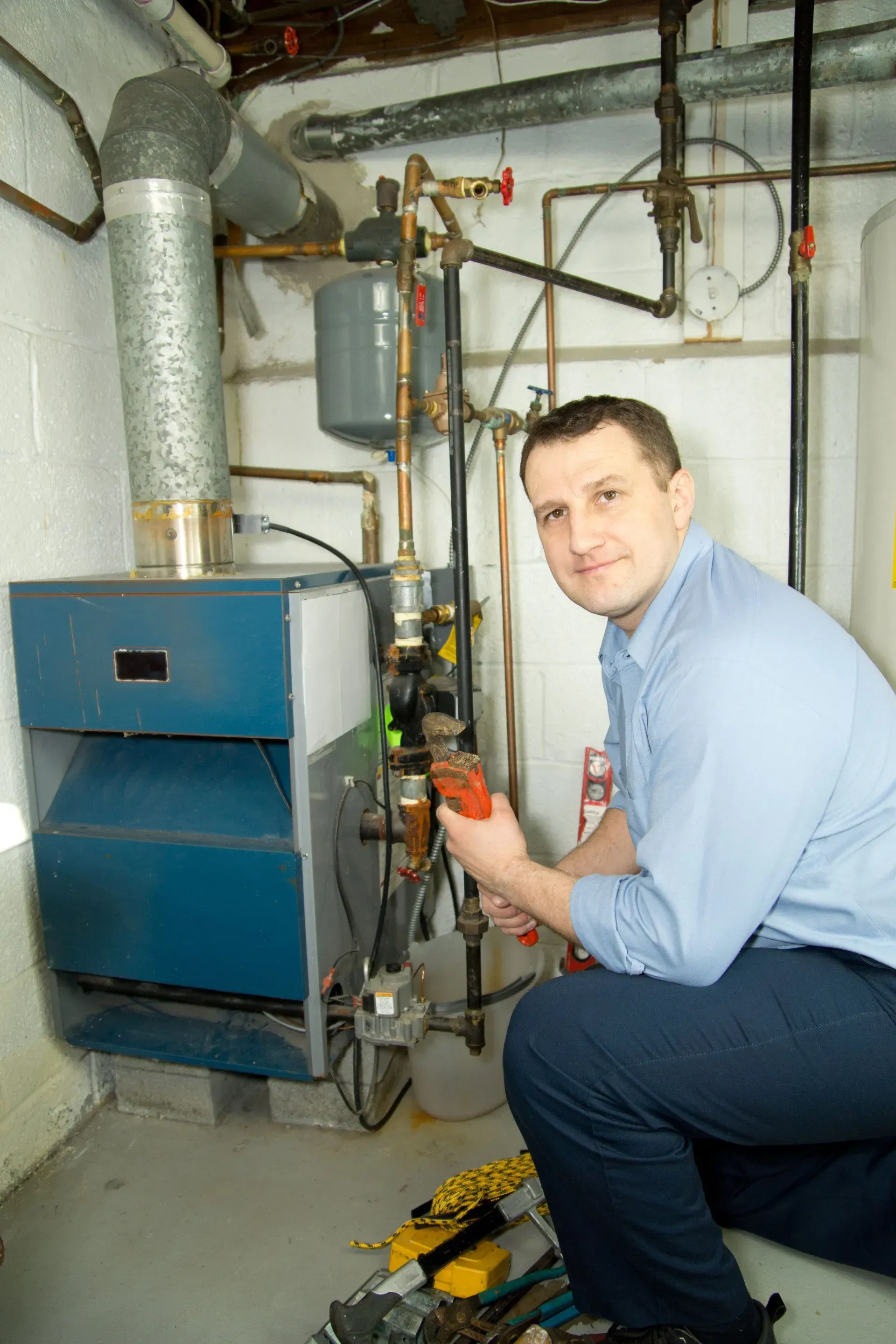 A professional kneeling to repair a blue boiler in a basement, holding a tool near the pipe connections.