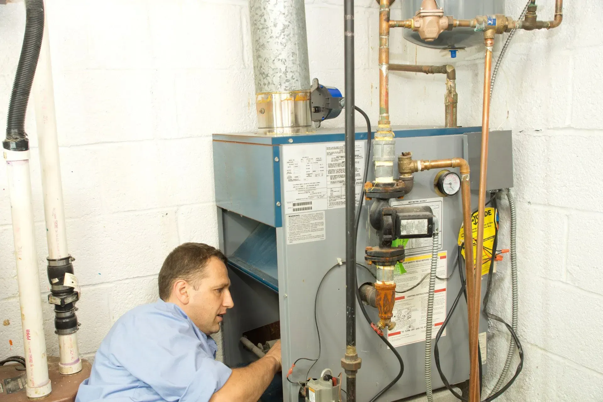 A technician in a blue shirt repairs a residential boiler system in a basement with white concrete block walls.