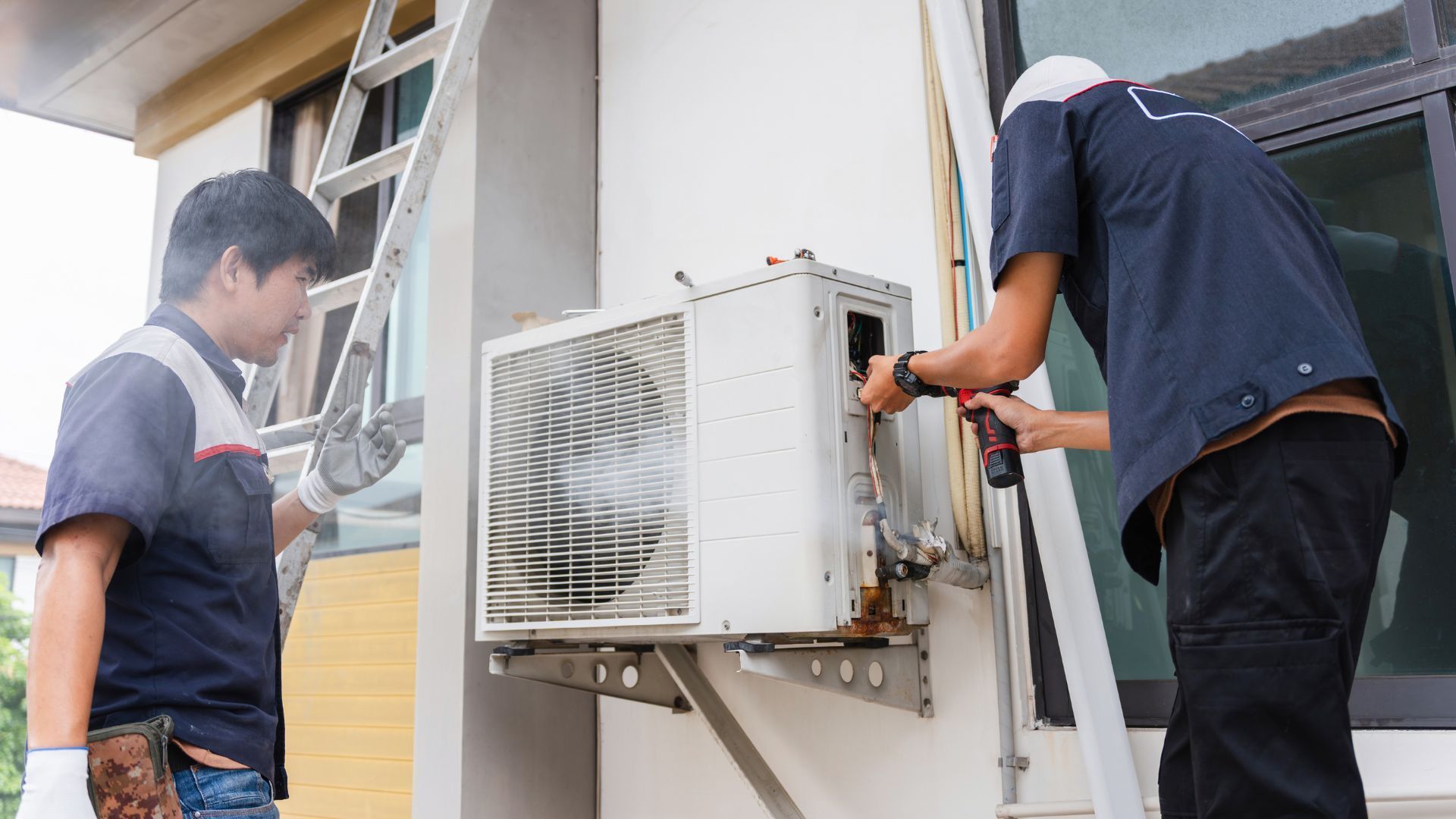 Two workers in uniforms repair an air conditioning condenser unit mounted on an exterior wall.
