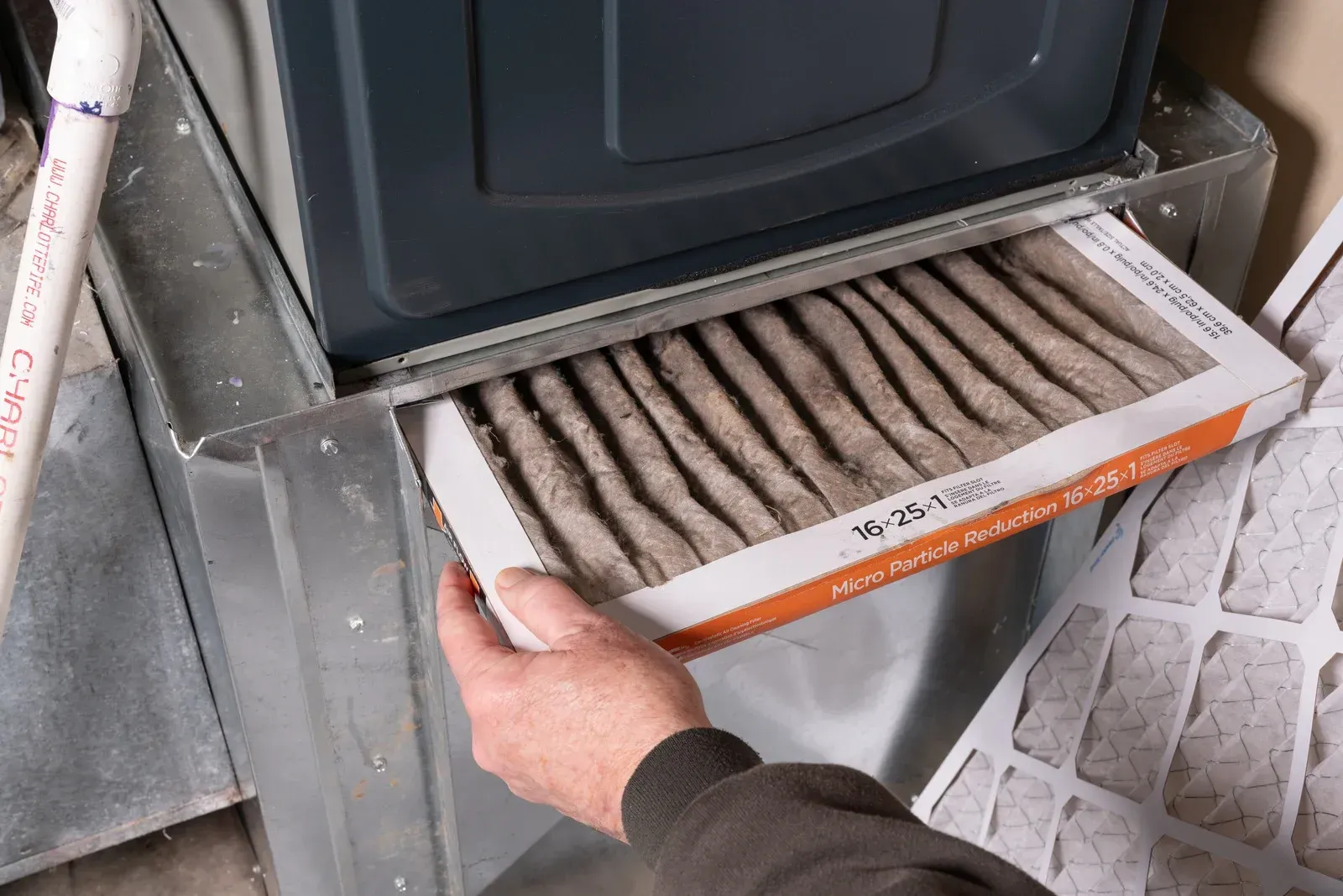 A hand pulling a dirty, dust-covered furnace air filter from a metal housing unit to replace it with a clean one.