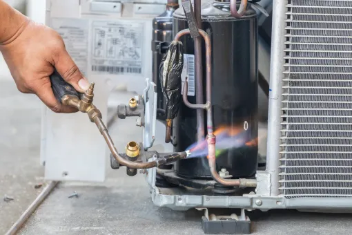A technician uses a torch to braze or solder a copper refrigerant pipe on an outdoor air conditioning unit.
