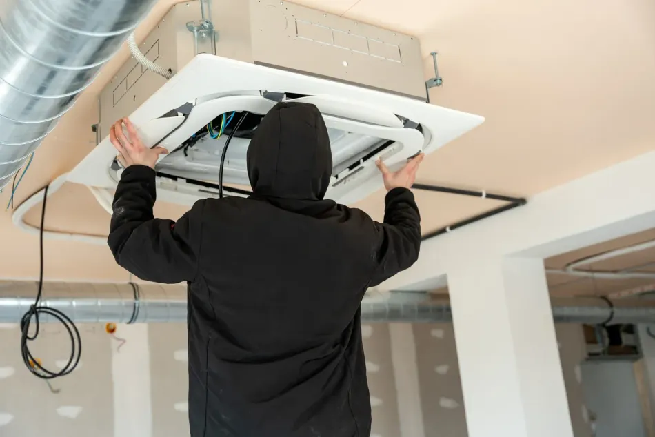 A person in a black hooded sweatshirt installing a white ceiling-mounted HVAC unit in an unfinished room.