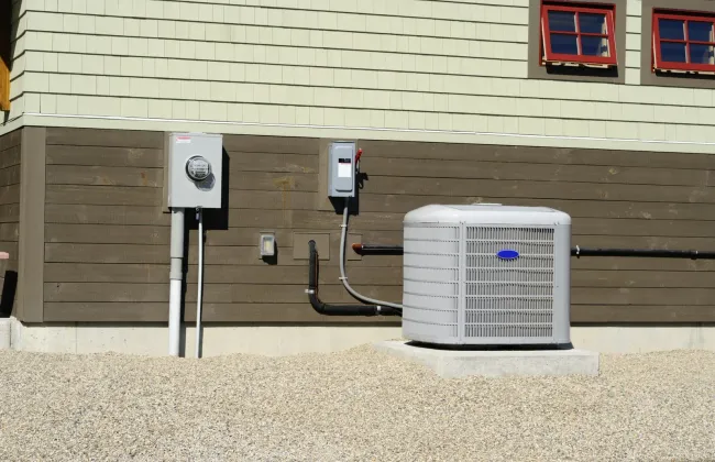 Exterior of a house showing a grey central air conditioning unit, electrical meter, and disconnect box on a gravel bed.