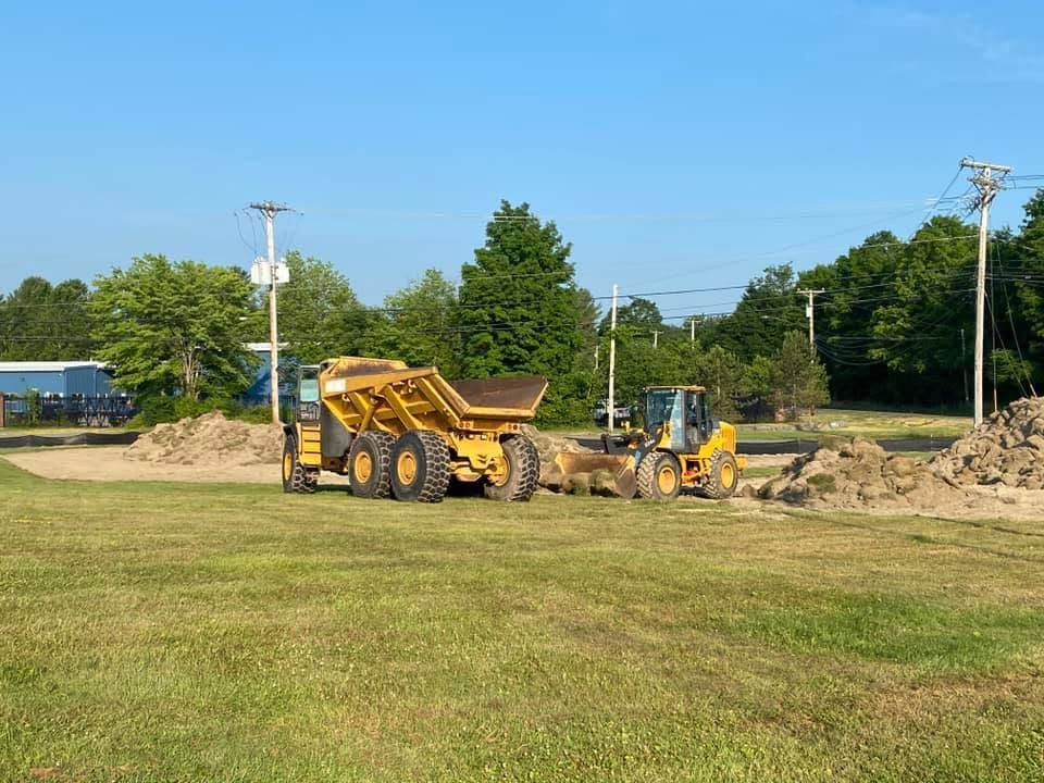 Construction equipment working on a dirt pile in a grassy field under a clear blue sky