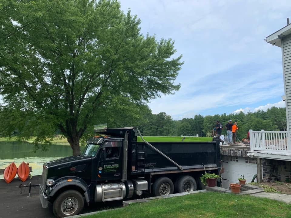 Black dump truck parked beside a house, with workers on the driveway and a pond in the background
