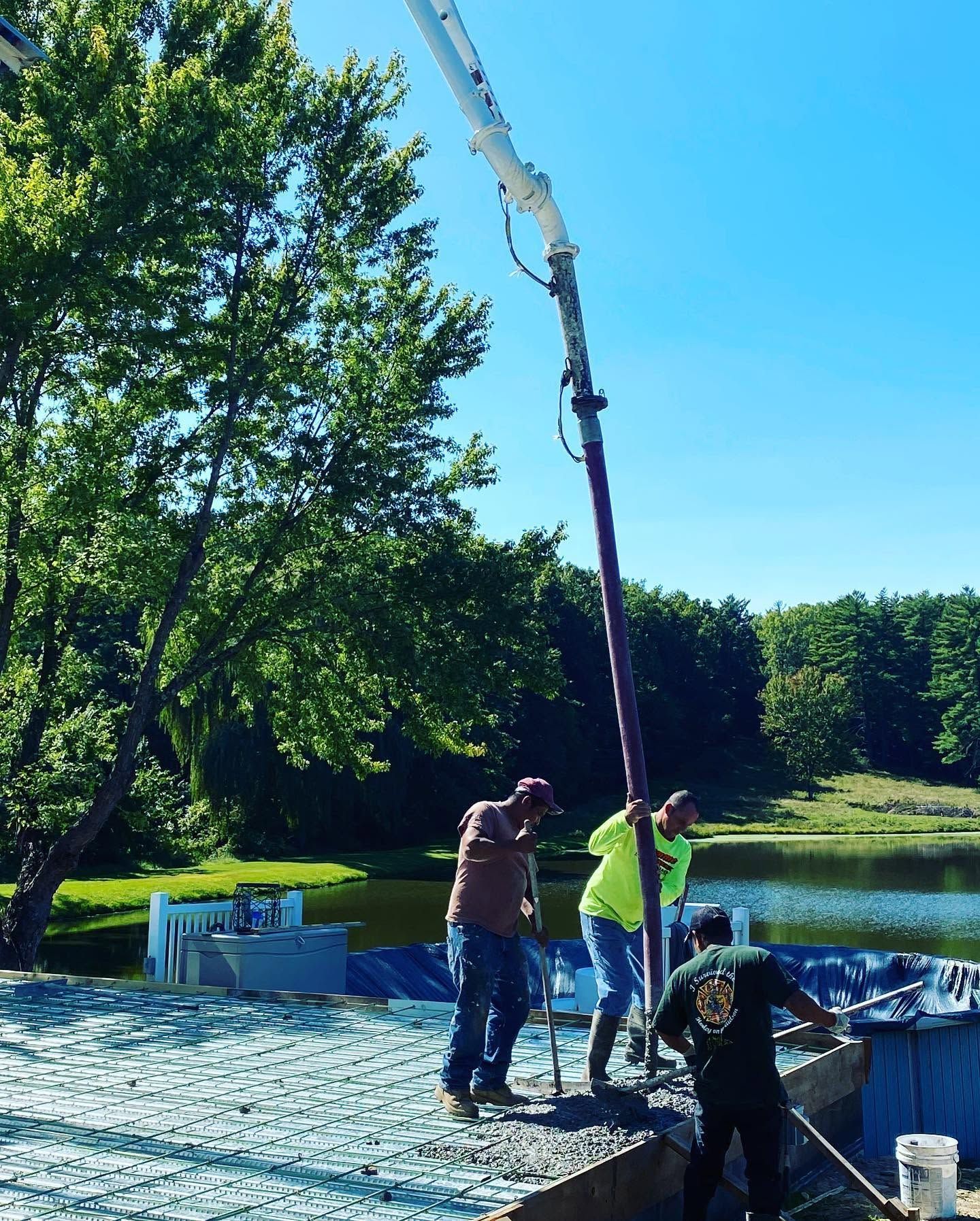Workers use a crane to lift a long pole beside a pond on a sunny day