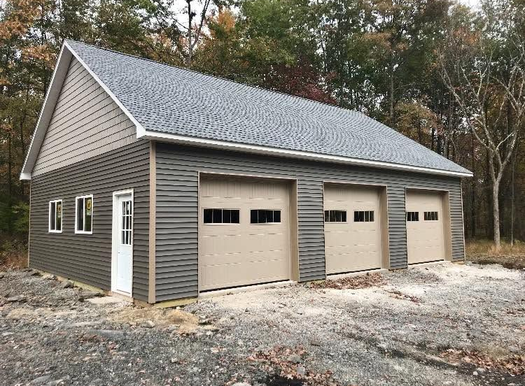 Detached gray three-car garage with beige doors and white trim beside trees