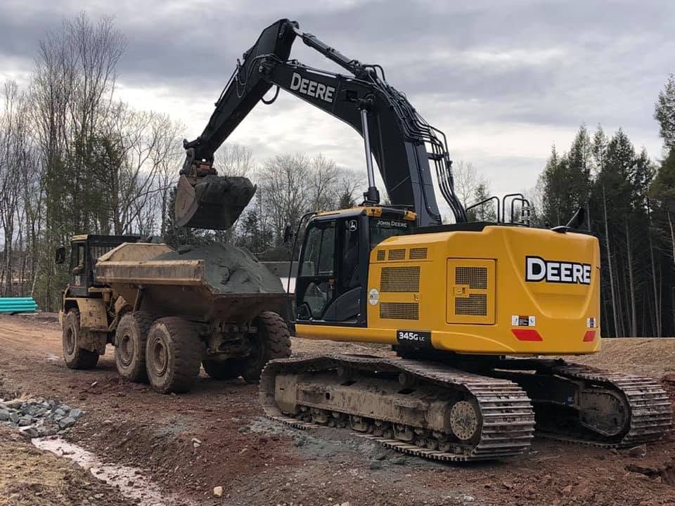 Yellow John Deere excavator dumping dirt into a dump truck on a muddy construction site.