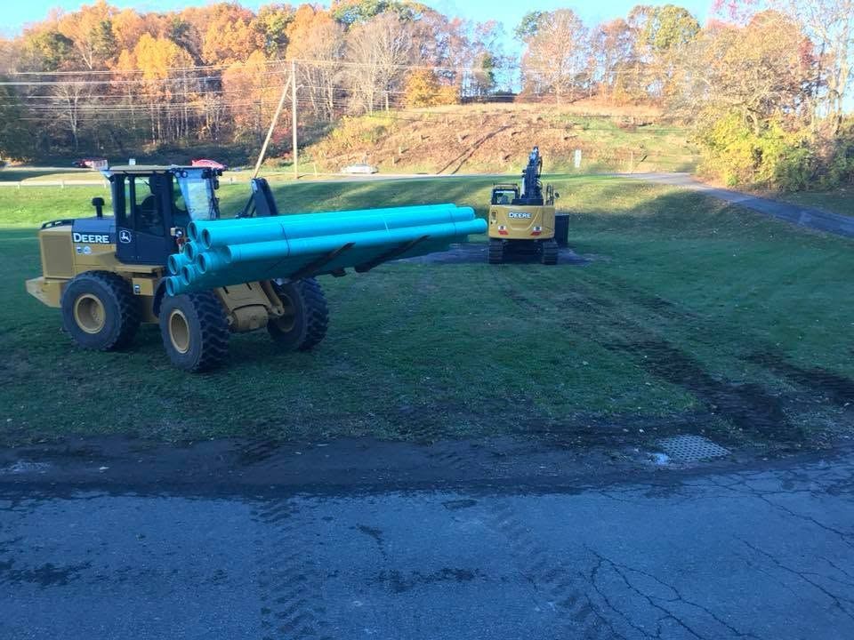 Two bulldozers hauling long blue pipes across a grassy field near a road in autumn.