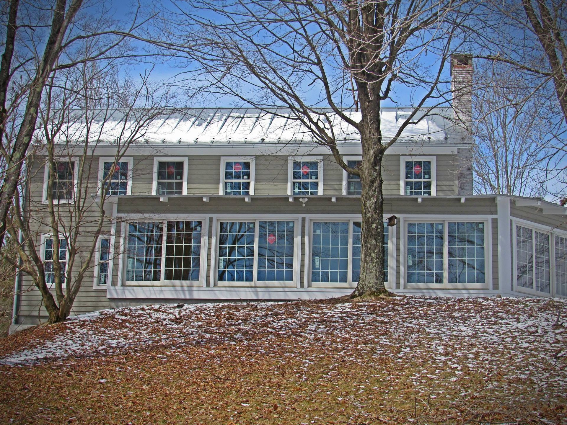 Snow-dusted two-story house with large windows and leafless trees in front, under a clear blue sky