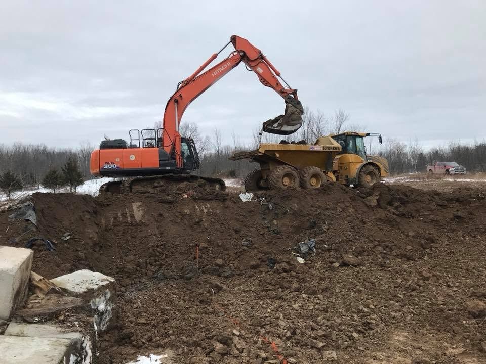 Orange excavator loading dirt into a yellow dump truck on a muddy construction site under cloudy sky