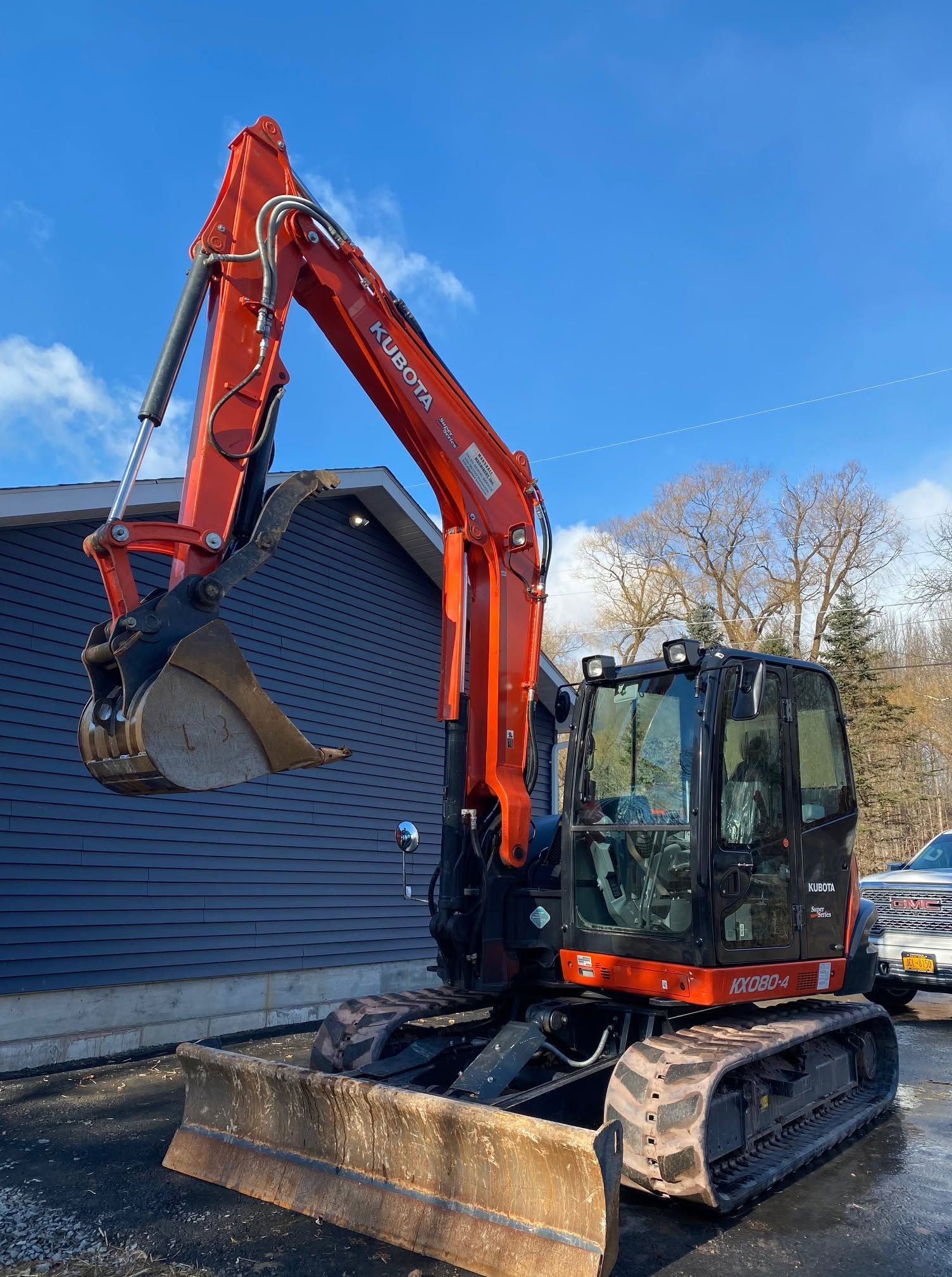 Orange excavator lifting a large round wooden object beside a house under a blue sky
