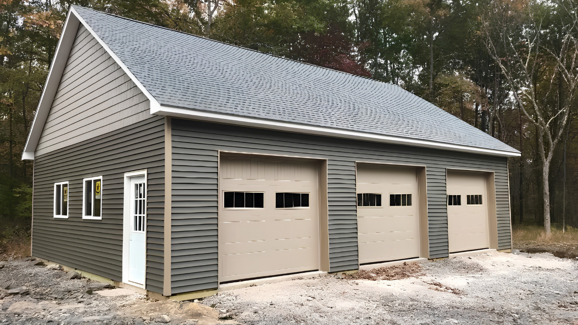 Detached three-car garage with gray siding and beige doors, shown on a gravel driveway in a wooded area