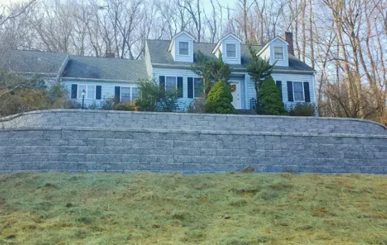 White house with blue shutters and dormers behind a stone retaining wall on a wooded hillside