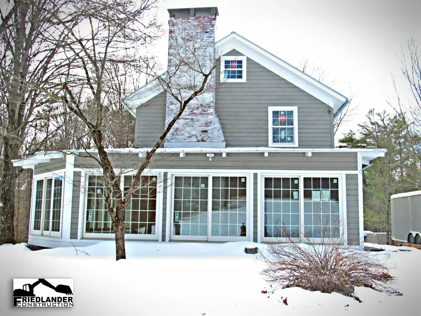 Snow-covered gray house with a stone chimney, large windows, and a screened porch