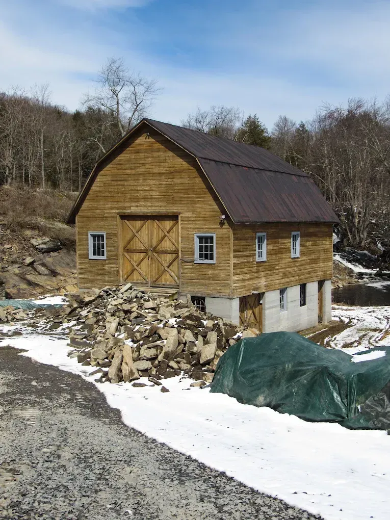 Wooden barn under construction beside a gravel road, with snow, stone piles, and a green tarp.