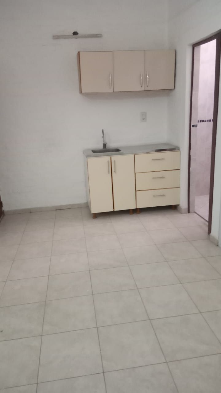 Pequeña cocina con armarios de color claro, fregadero y suelo de baldosas. Se ve una puerta a la derecha.