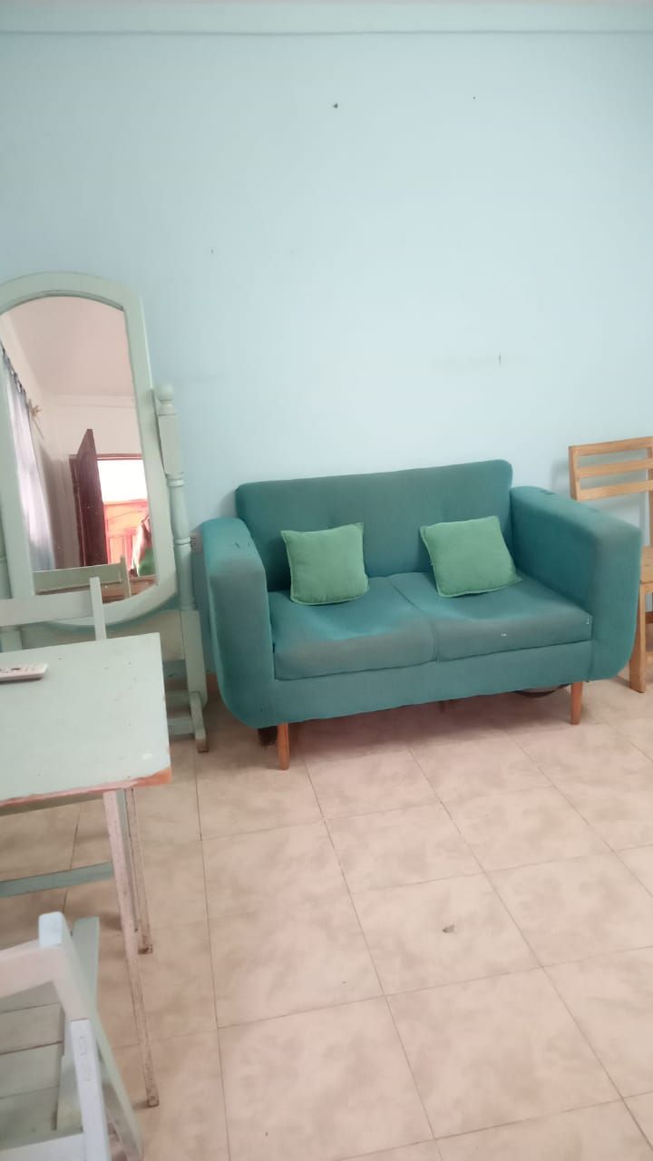 Sofá de dos plazas de color verde azulado con dos almohadas verdes contra una pared azul claro, un espejo y una mesa pequeña.