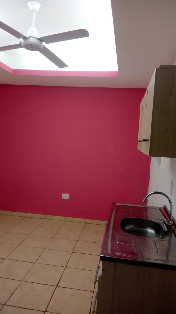 Una cocina pequeña con una pared rosa, ventilador de techo y fregadero. Hay armarios y suelo de baldosas.