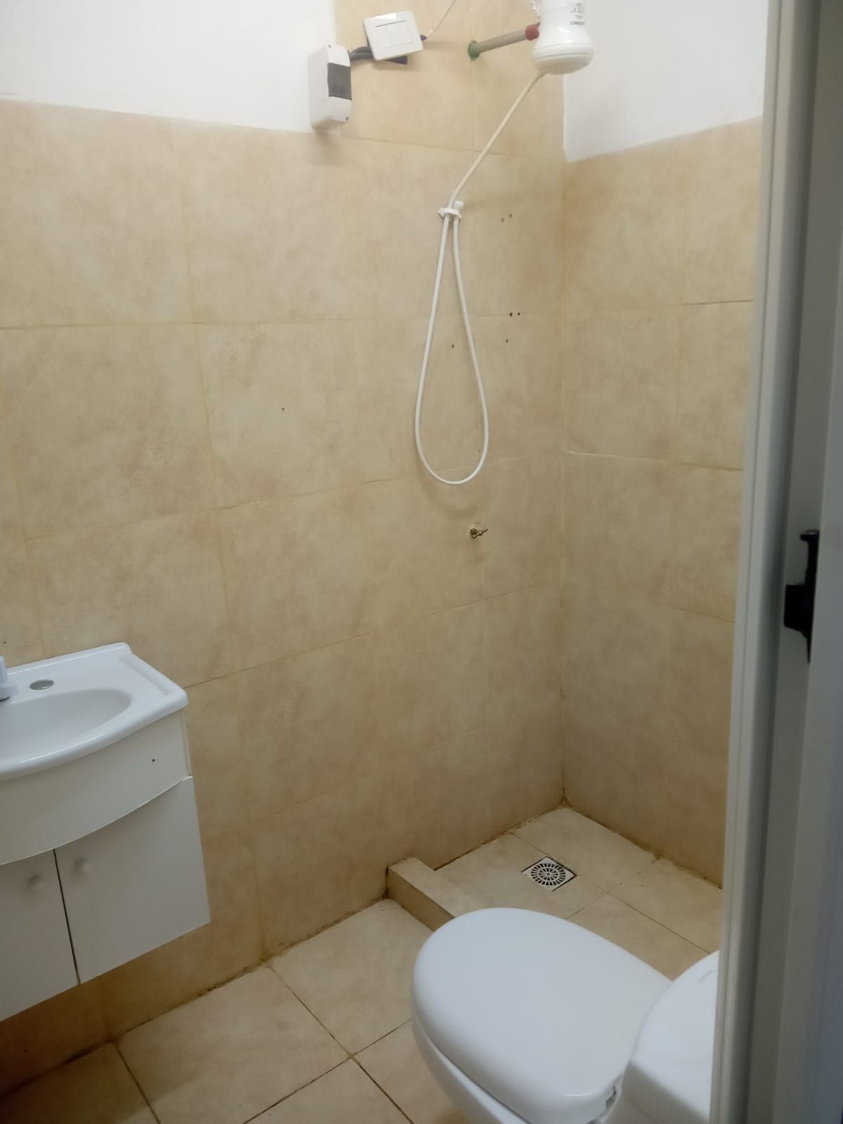 Baño pequeño con inodoro, lavabo y ducha. Paredes y suelo de azulejos beige; inodoro y lavabo blancos.